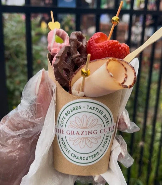 Hand holding a cup of food with a 'The Grazing Cutie' label, against a blurred background.