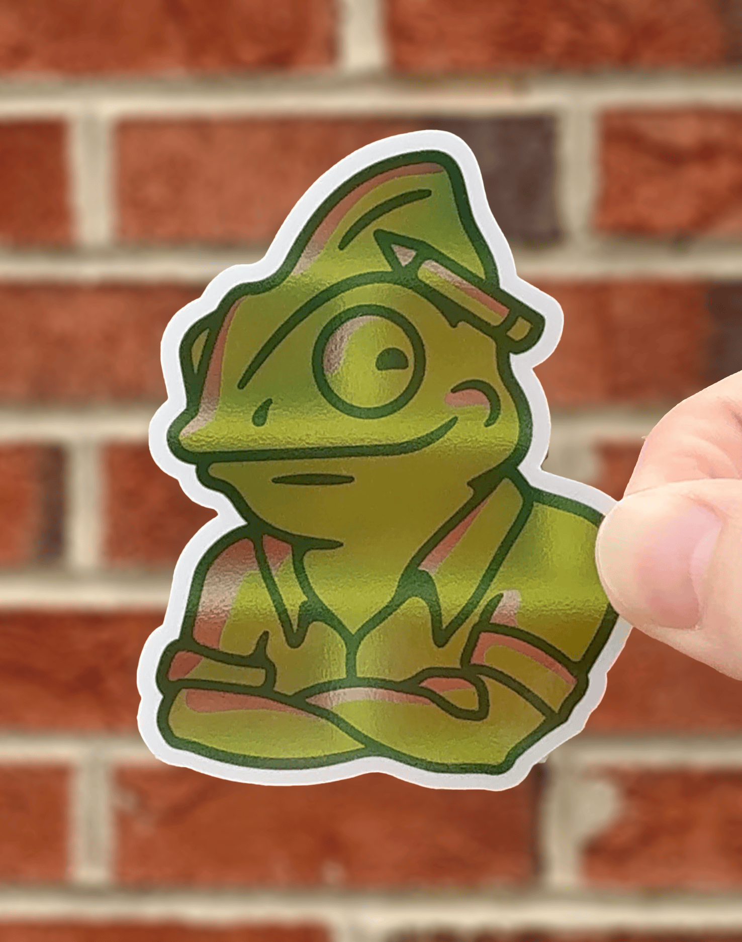 Green cartoon lizard sticker held against a brick wall background