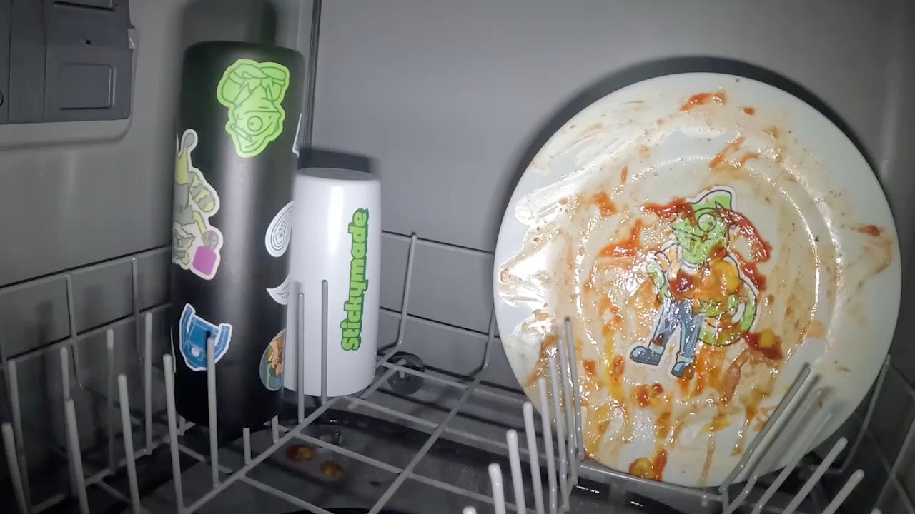 A dirty plate with a sticker on it, a bottle and a cup covered in stickers, going into the dishwasher